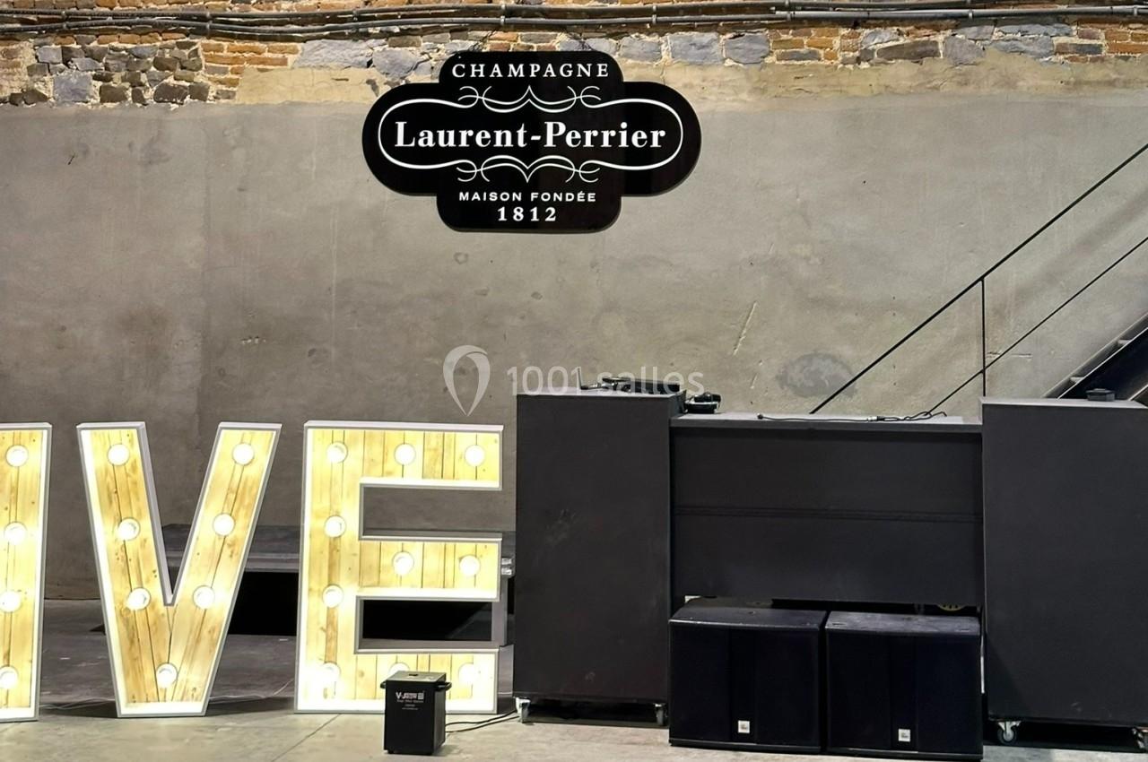 Panneau mural ’Laurent-Perrier’ avec des lettres lumineuses ’LIVE’ et un équipement audio devant un mur en briques.