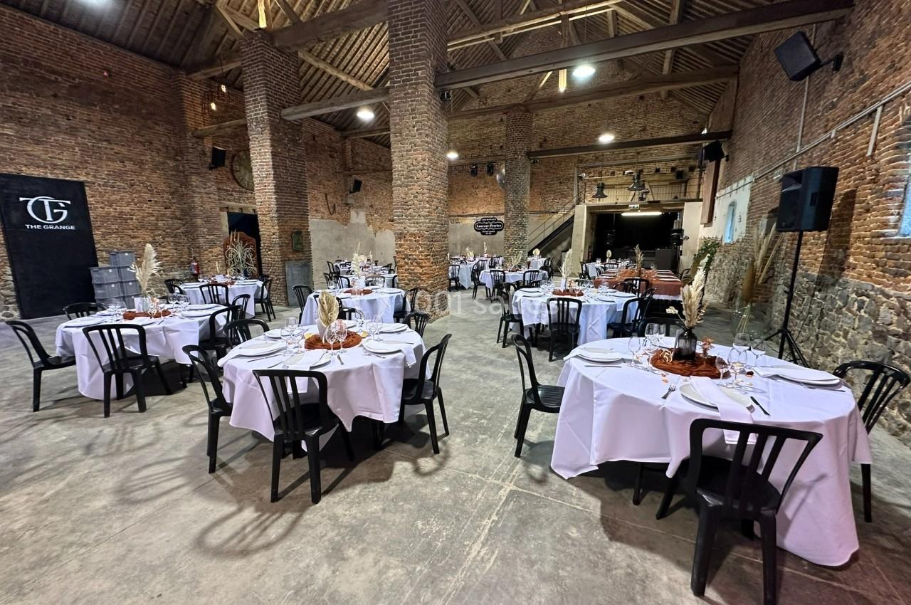 Salle en briques avec plafond en poutres apparentes, tables rondes dressées avec nappes blanches et chaises noires.