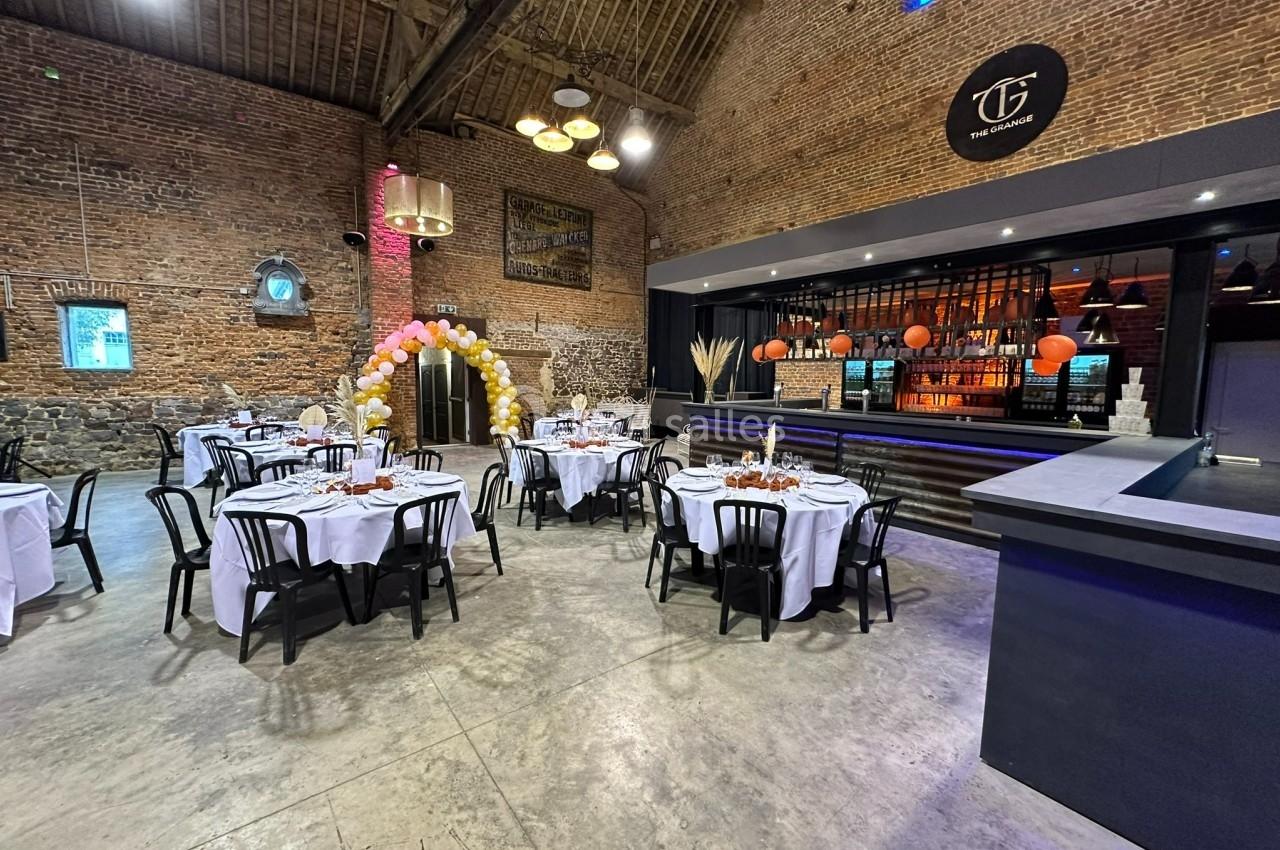 Salle de réception avec tables dressées, arche de ballons, murs en briques et bar éclairé en arrière-plan.