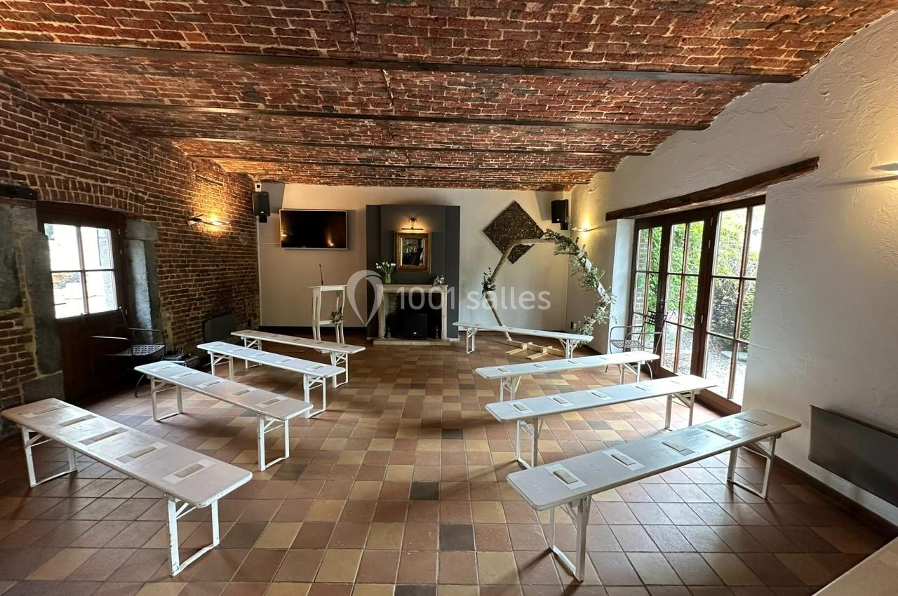 Salle en briques avec plafond voûté, bancs blancs disposés en rangées, cheminée et grande baie vitrée donnant sur un jardin.