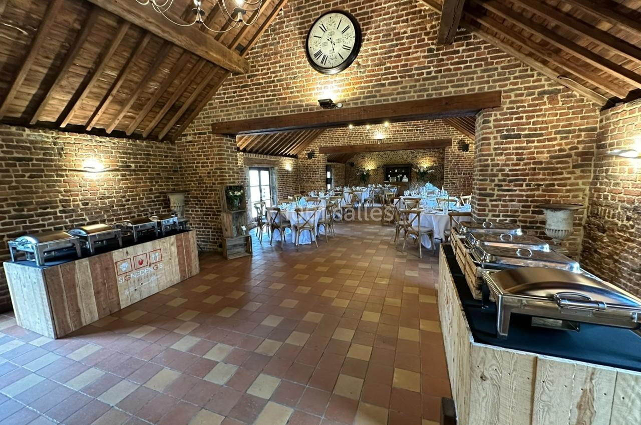 Salle de réception en briques avec tables dressées, buffet équipé et grande horloge murale.