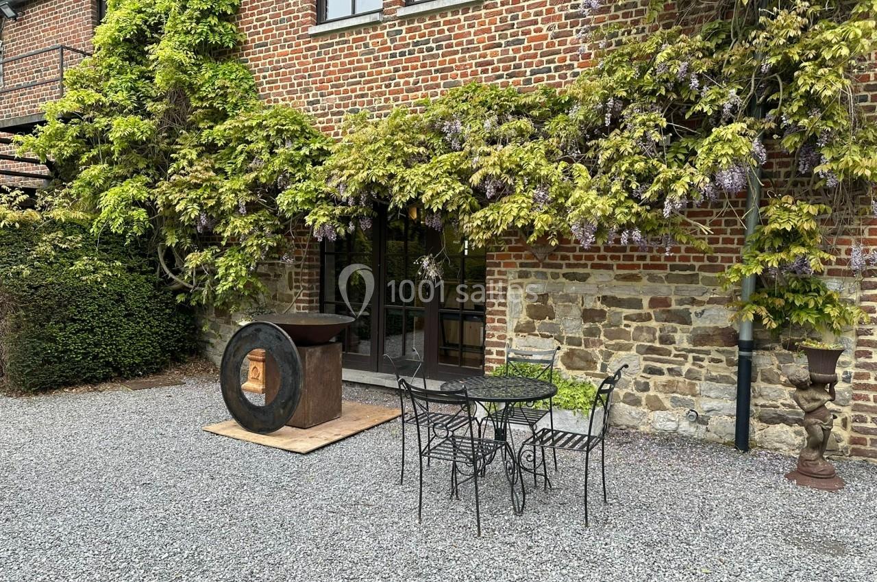 Façade en briques avec vigne grimpante, table et chaises en fer forgé sur une cour gravillonnée.