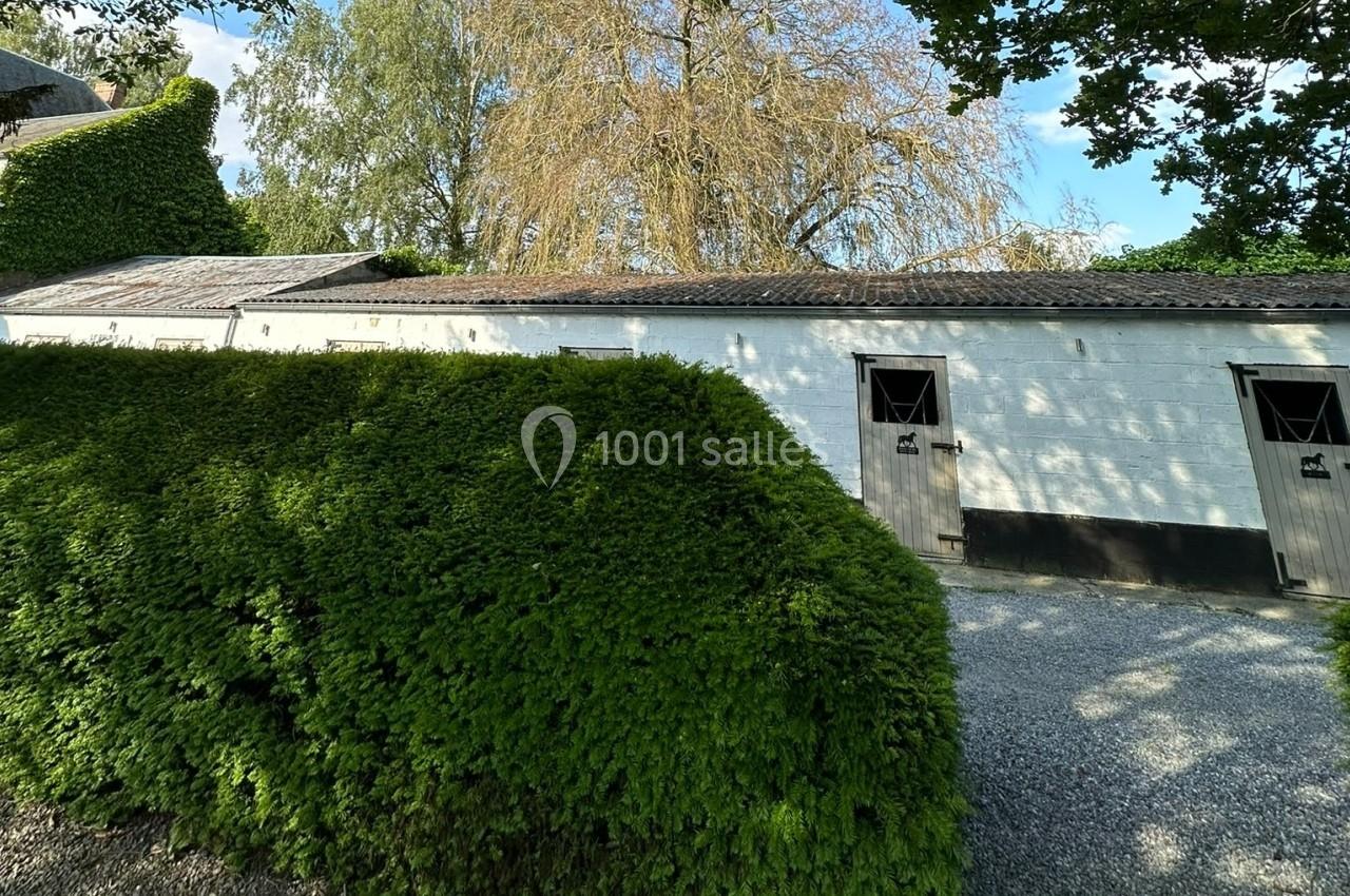 Bâtiment blanc avec portes en bois, entouré de haies taillées et d'arbres, sous un ciel partiellement dégagé.