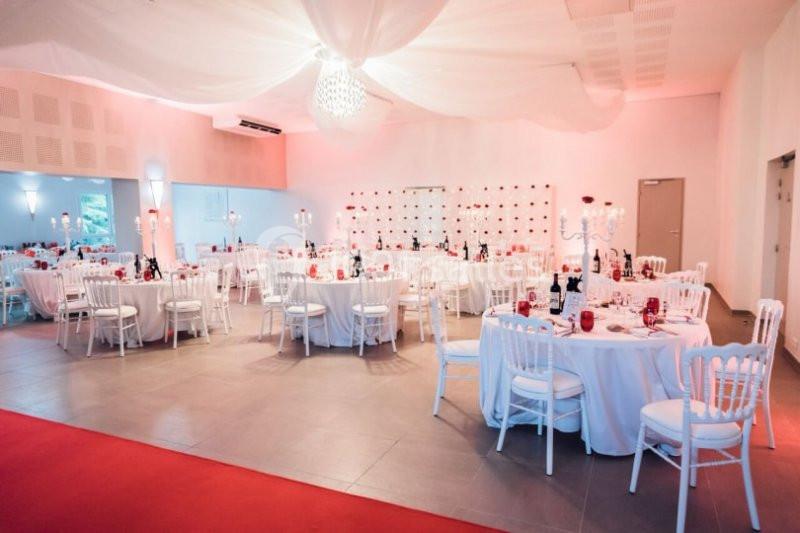 Salle de réception décorée avec des tables rondes dressées, chaises blanches et éclairage tamisé rose.