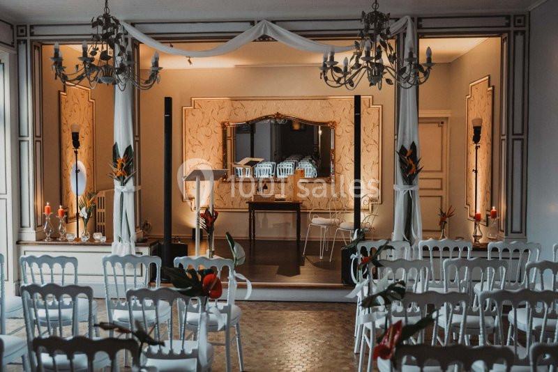 Salle décorée pour une cérémonie, avec des chaises blanches alignées, des fleurs tropicales et un espace central aménagé.