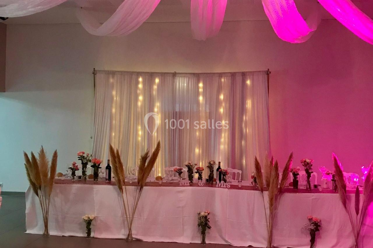 Décoration de salle avec une table longue ornée de fleurs, pampas et guirlandes lumineuses sous un éclairage rose.