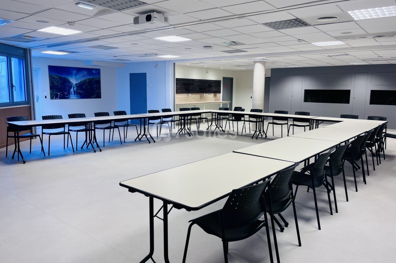 Salle de réunion spacieuse avec tables disposées en U, chaises noires et éclairage naturel provenant d'une fenêtre.