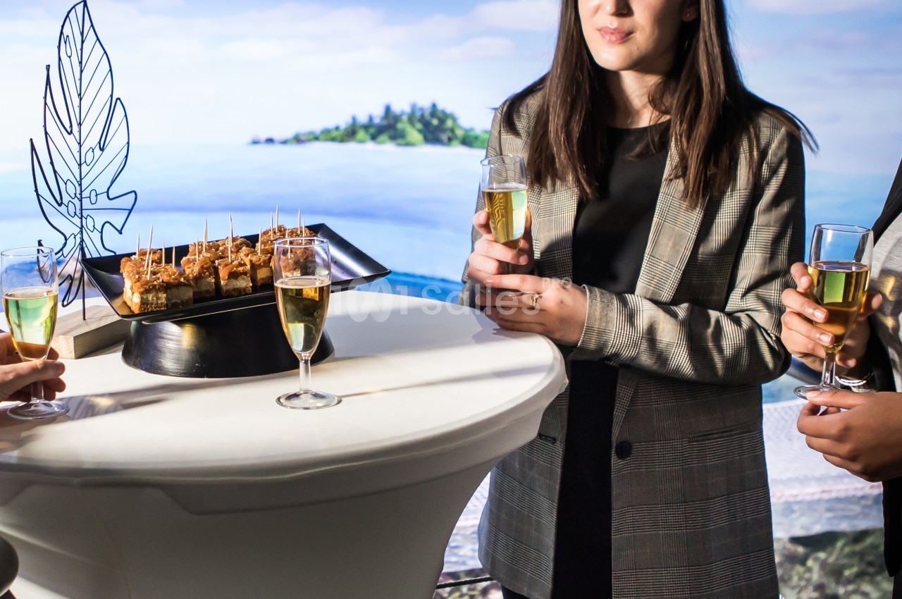 Trois personnes debout autour d'une table haute avec des amuse-bouches et des verres de champagne, en arrière-plan un…