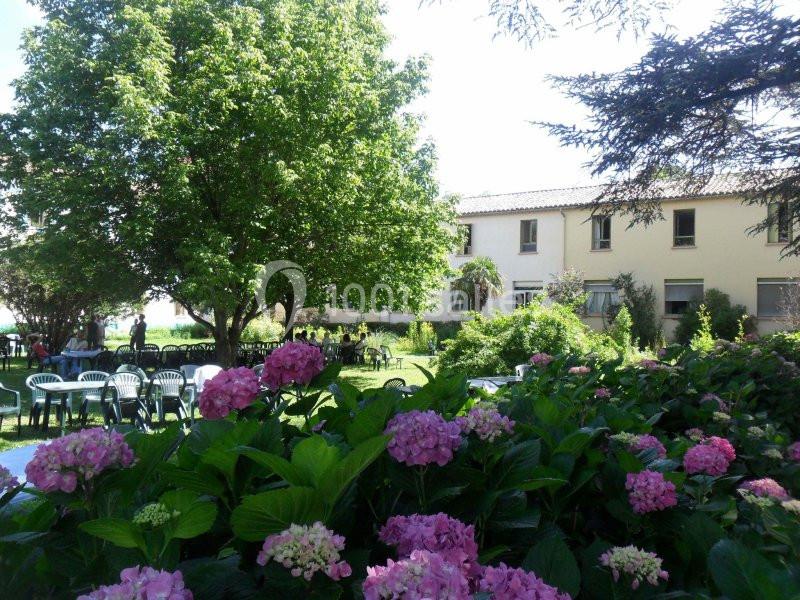 Jardin verdoyant avec des hortensias roses au premier plan, des arbres et un bâtiment beige en arrière-plan.