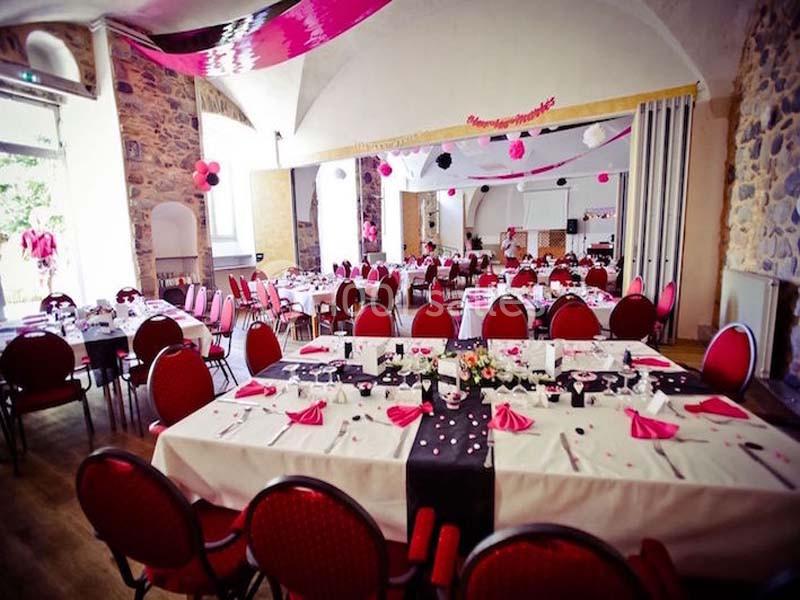 Salle décorée pour un événement avec des tables dressées, nappes blanches, serviettes roses et guirlandes suspendues.