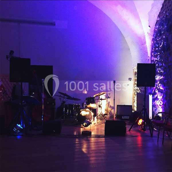 Scène de concert vide avec batterie, amplificateurs et éclairage coloré dans une salle voûtée.