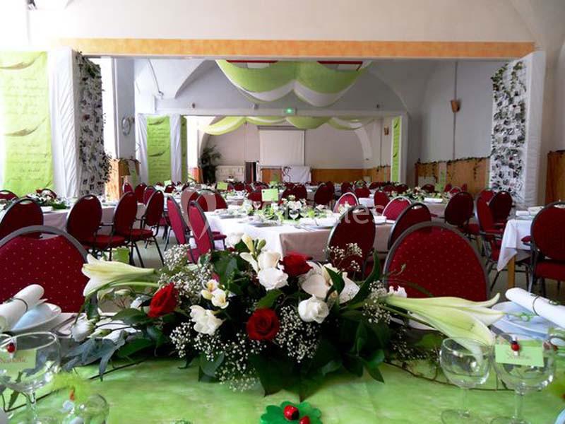 Salle décorée pour un événement avec des tables dressées, nappes vertes, chaises rouges et arrangements floraux.