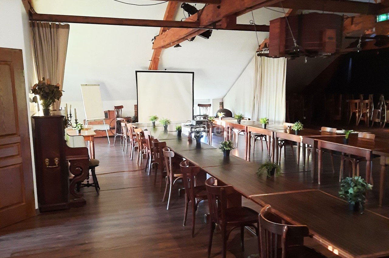 Salle de réunion lumineuse avec de longues tables en bois, des chaises, un écran de projection et des plantes décoratives.