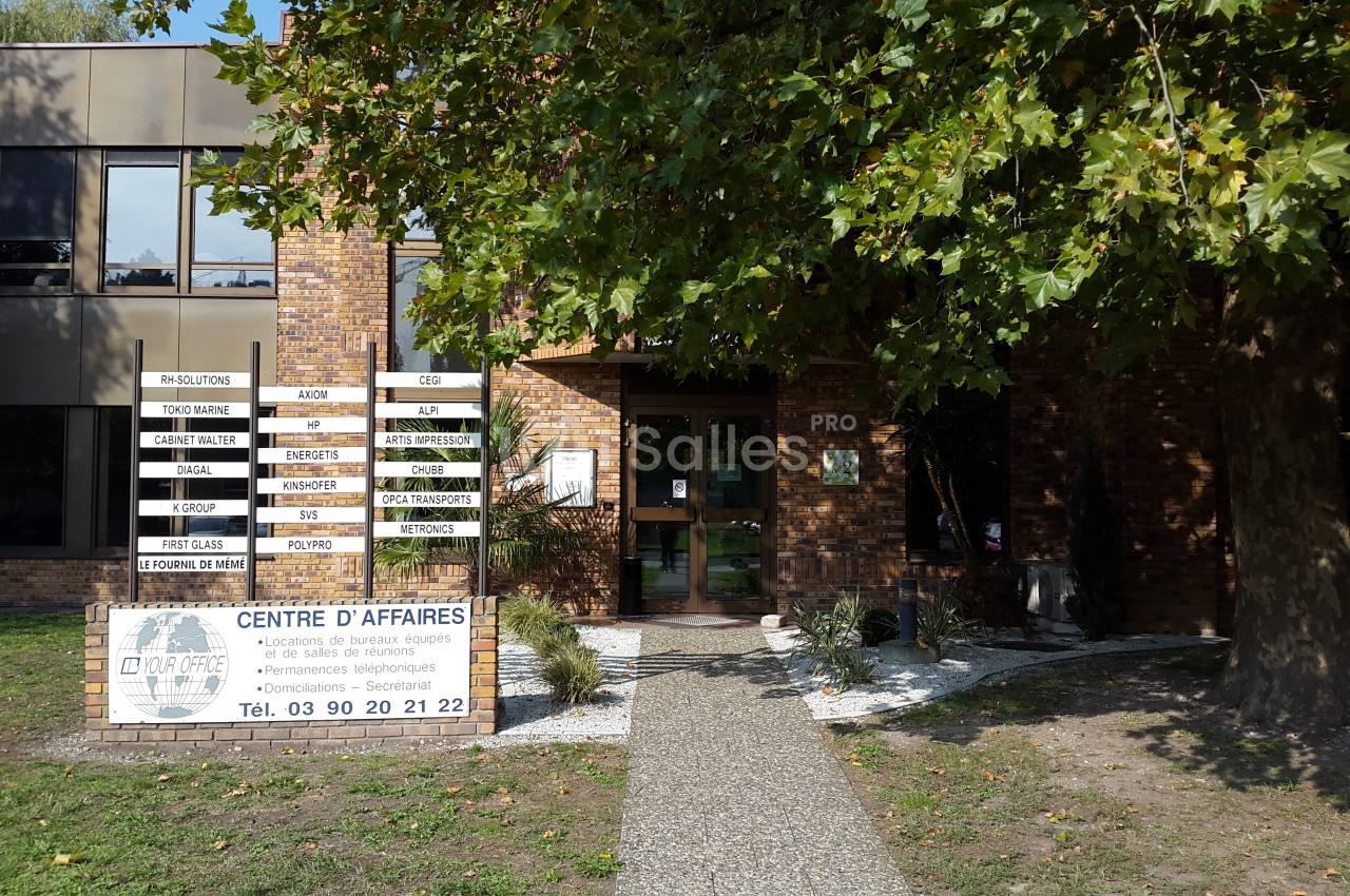 Façade d'un centre d'affaires en briques, avec une entrée bordée de végétation et des panneaux d'information.