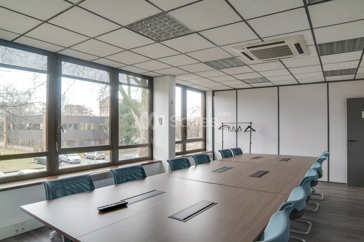 Salle de réunion lumineuse avec grande table en bois, chaises bleues, fenêtres donnant sur un extérieur arboré.