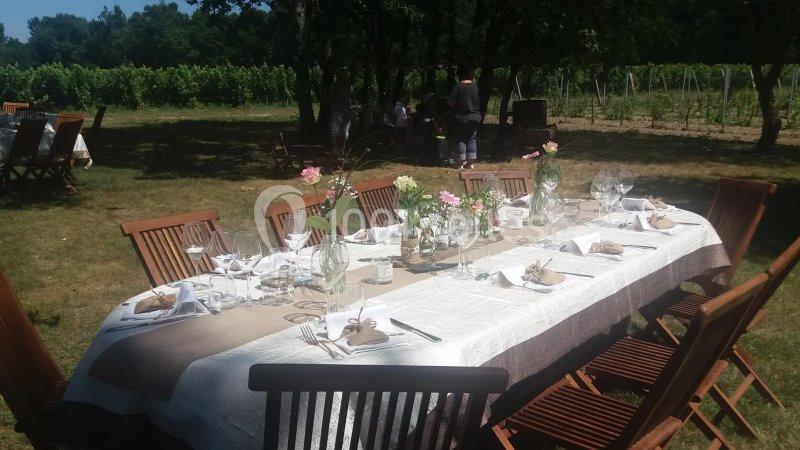 Table dressée en extérieur avec nappes, verres et décorations florales, entourée de chaises en bois, près de vignes.