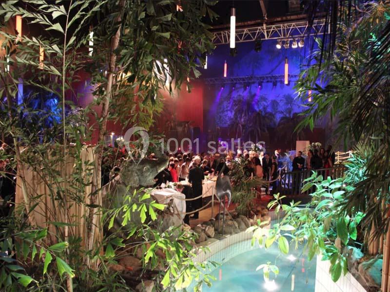 Salle événementielle décorée de plantes exotiques, avec un bassin au premier plan et une foule rassemblée en arrière-plan.