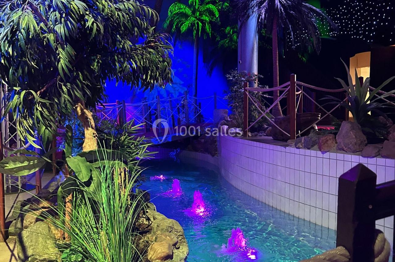 Cours d'eau artificiel entouré de plantes tropicales et éclairé par des lumières colorées dans un espace intérieur.