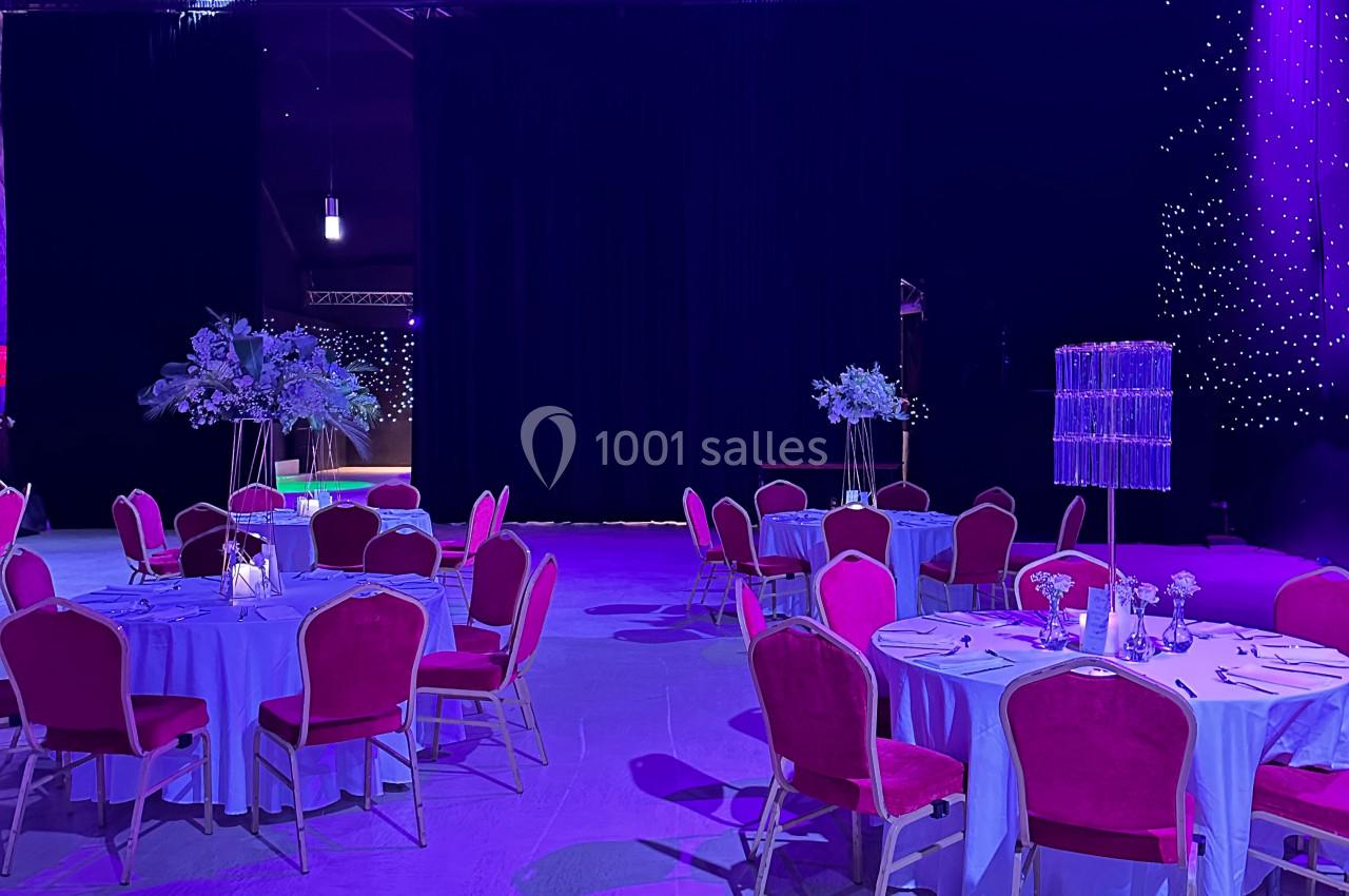 Salle de réception décorée avec des tables rondes, nappes blanches, chaises rouges et éclairage violet.