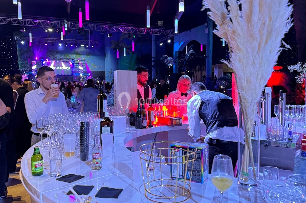 Bar lumineux en forme de cercle avec verres et bouteilles, dans une salle animée éclairée par des lumières colorées.