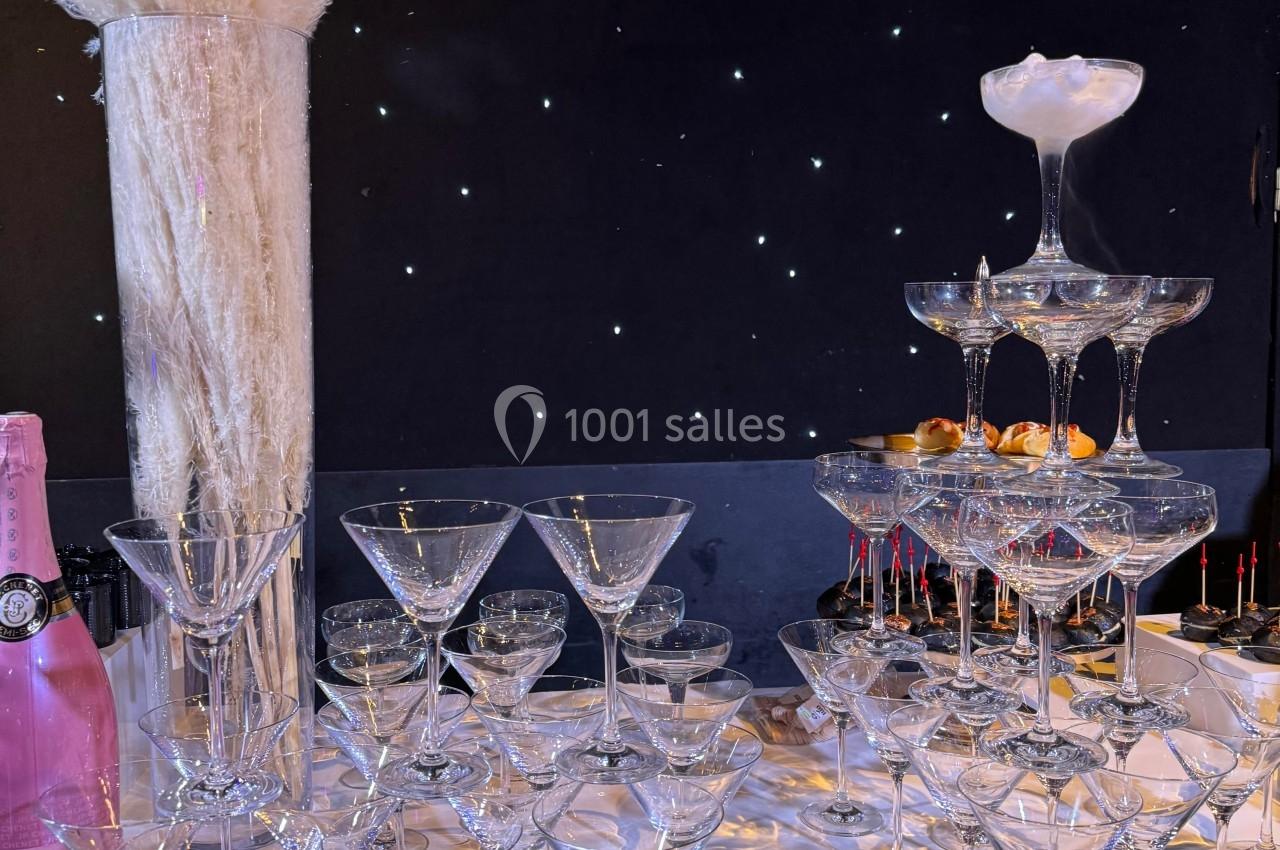 Table décorée avec des verres à cocktail empilés en pyramide, un vase de pampas et une bouteille rose, sur fond étoilé.