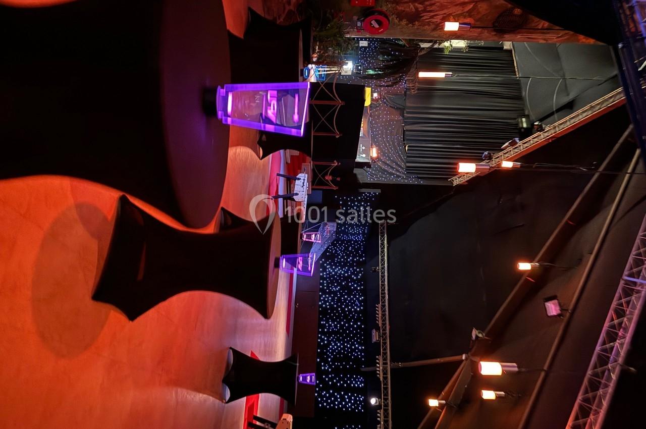 Salle sombre avec tables hautes noires, éclairage tamisé et décorations lumineuses en arrière-plan.