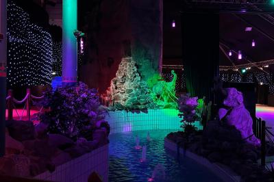 Décor intérieur avec éclairage coloré, fontaines illuminées, végétation tropicale et tables dressées en arrière-plan.