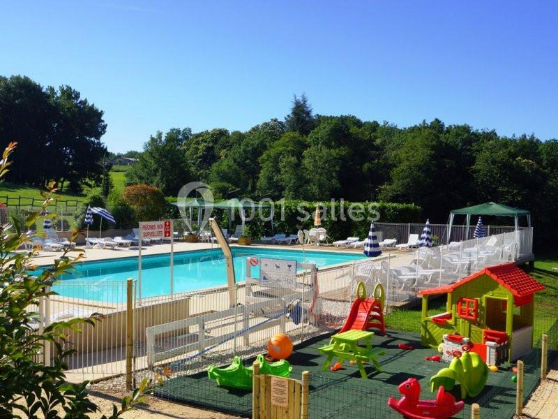 Piscine extérieure avec transats et parasols, aire de jeux pour enfants clôturée au premier plan, entourée de verdure.