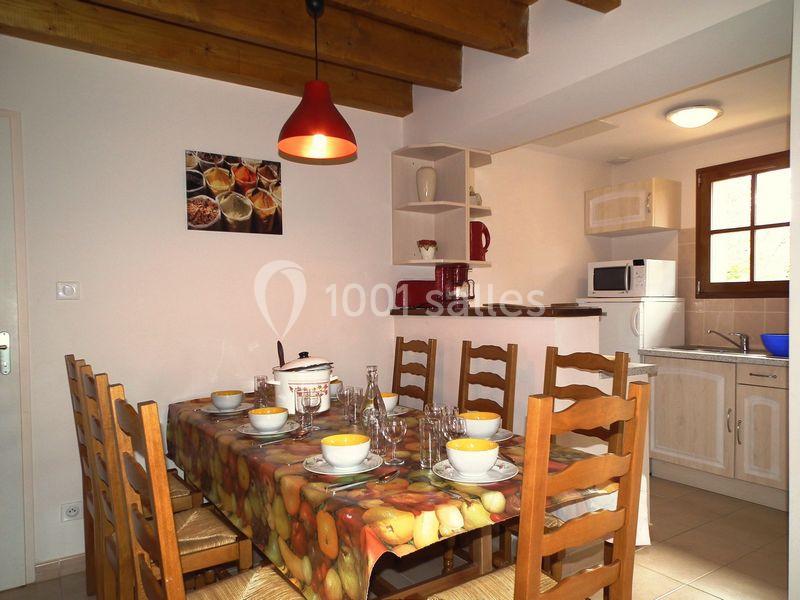 Salle à manger avec table dressée pour six personnes, cuisine ouverte équipée en arrière-plan.