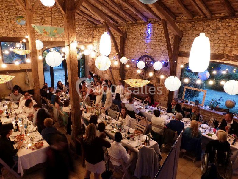 Salle en pierre avec des poutres apparentes, décorée de guirlandes lumineuses, accueillant un dîner festif avec de nombreux…