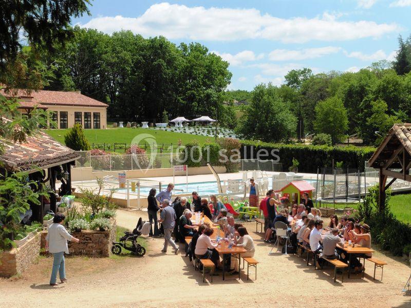 Des personnes déjeunent en plein air à des tables en bois près d'une piscine, entourées de verdure et de bâtiments.