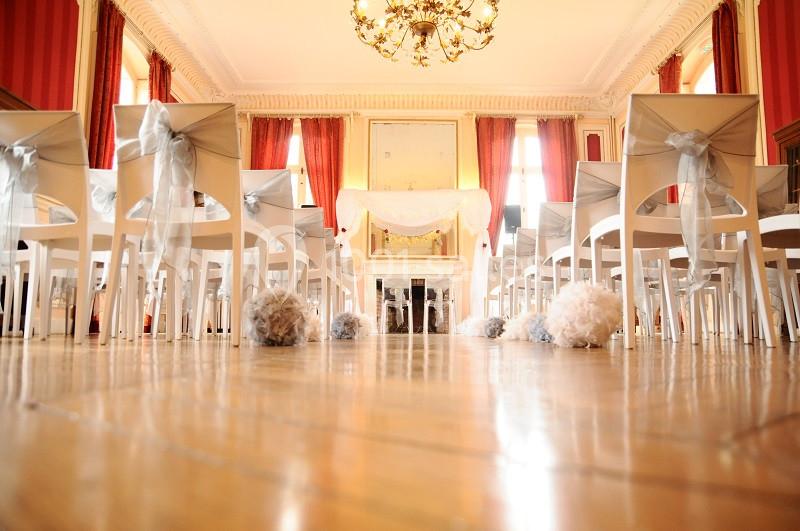 Salle de cérémonie décorée avec des chaises blanches ornées de rubans et un autel devant des rideaux rouges.