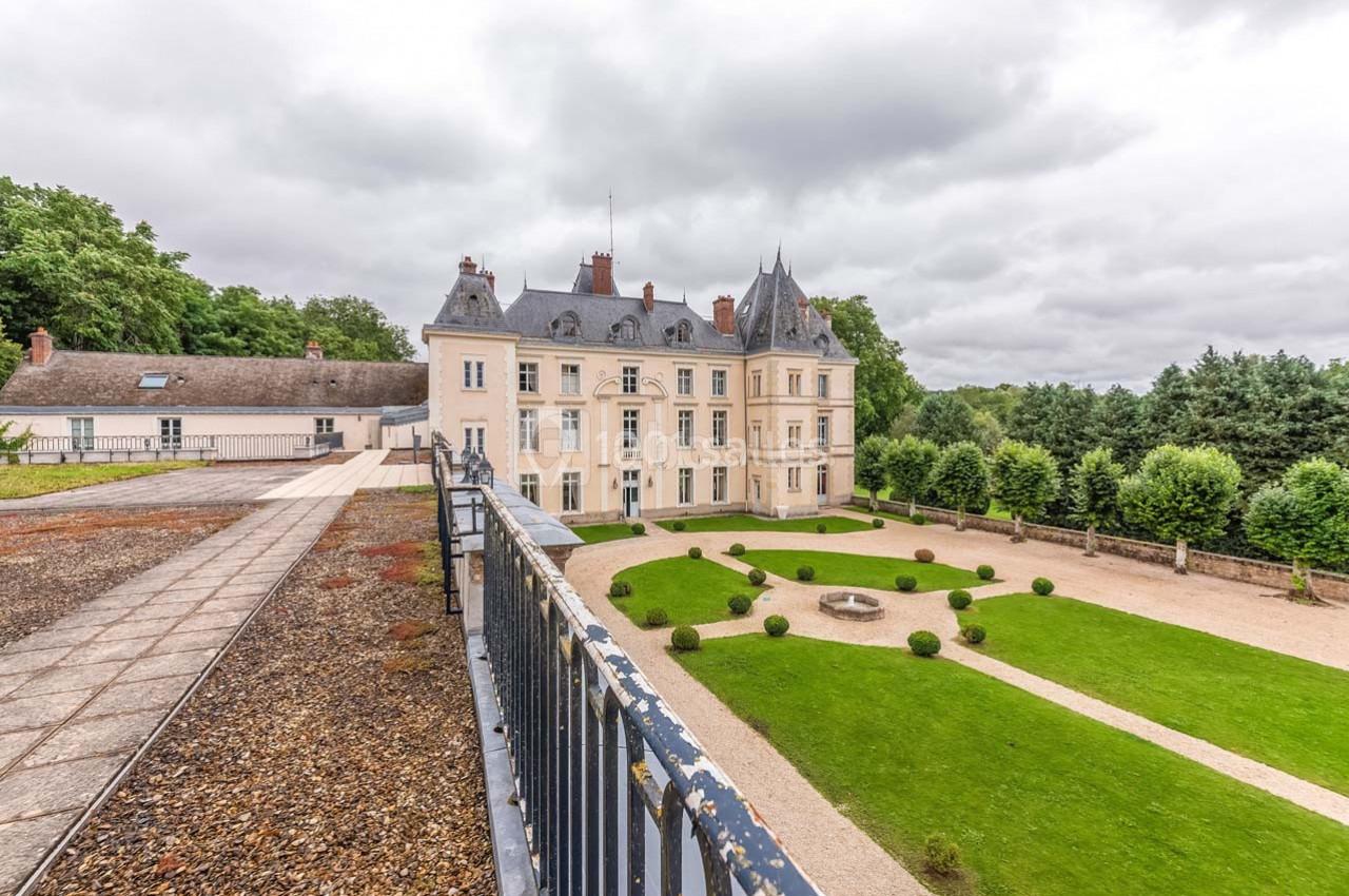 Façade d'un château entouré de jardins bien entretenus, avec un ciel nuageux en arrière-plan.