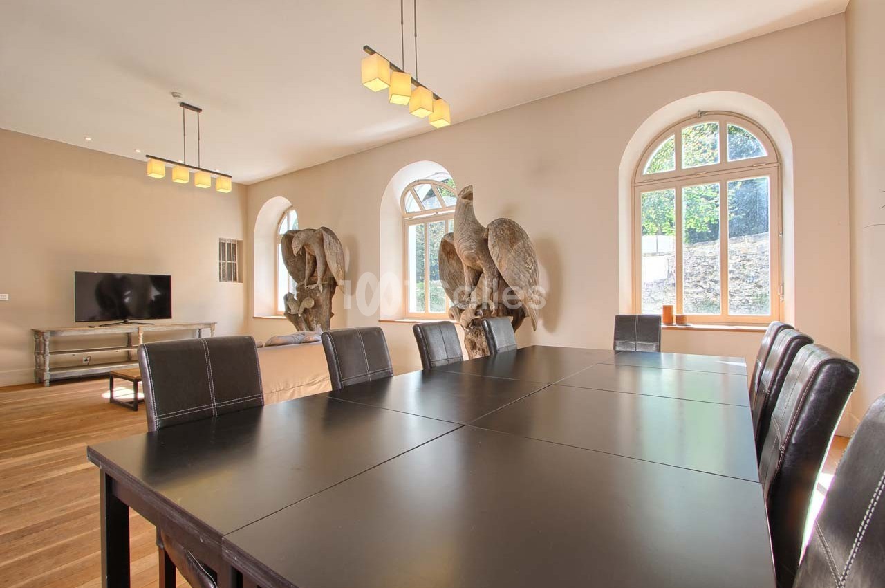 Salle à manger lumineuse avec une grande table noire, des chaises en cuir et des sculptures d'oiseaux près des fenêtres.