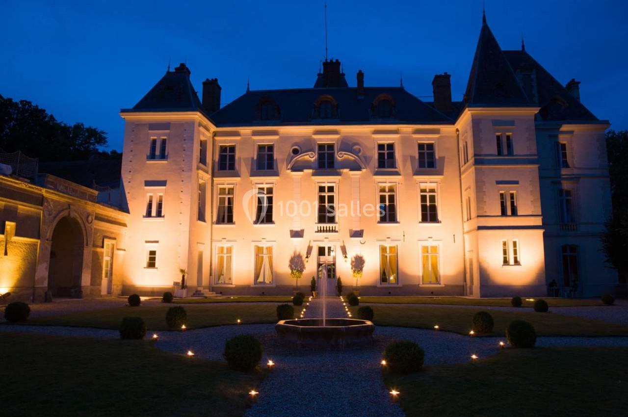 Façade d'un château illuminé de nuit, entouré d'un jardin avec une fontaine centrale et des allées éclairées.