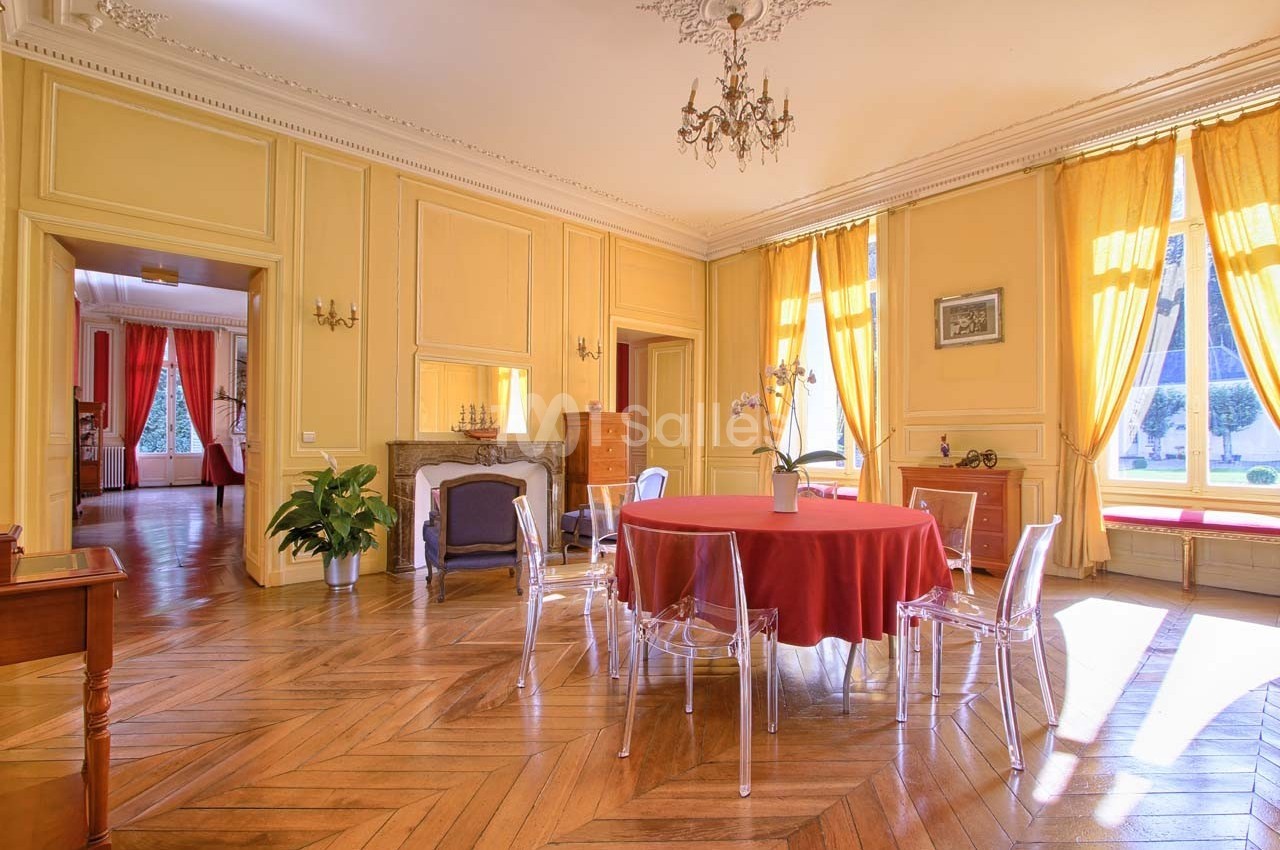 Salle lumineuse avec parquet en bois, murs jaunes, table ronde avec nappe rouge et chaises transparentes.