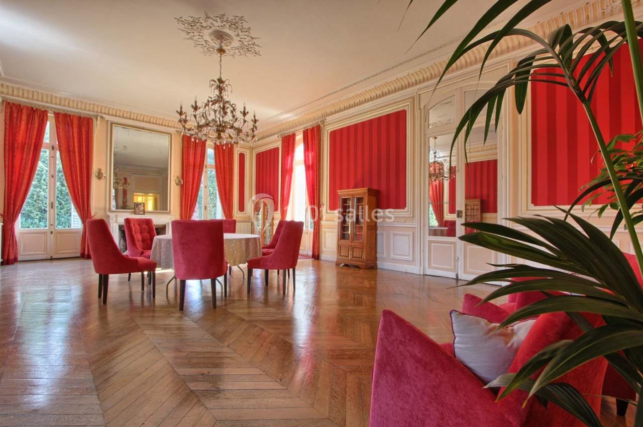 Salle élégante avec parquet en bois, murs rouges, rideaux assortis, lustre et mobilier en velours rouge.