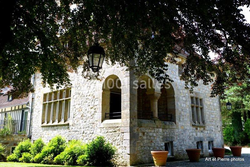 Maison en pierre avec arcades, fenêtres à carreaux et pots de fleurs, entourée de végétation et ombragée par un arbre.