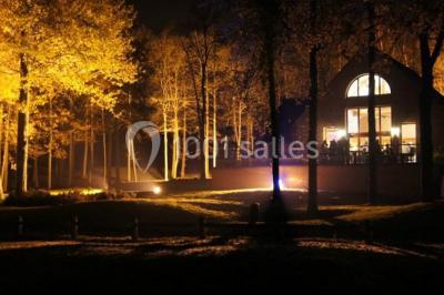 Vue nocturne d'une maison en bois éclairée, entourée d'arbres illuminés, sous un ciel nuageux avec la lune visible.