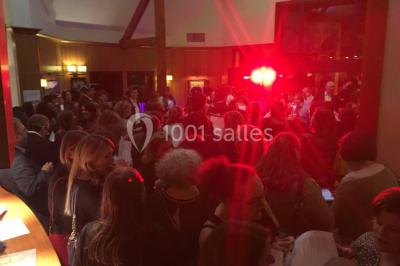 Vue nocturne d'une maison en bois éclairée, entourée d'arbres illuminés, sous un ciel nuageux avec la lune visible.