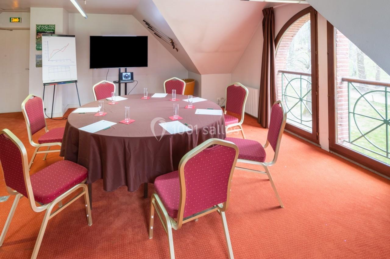 Salle de réunion lumineuse avec table ronde, chaises rouges, tableau blanc, écran et grandes fenêtres donnant sur un jardin.