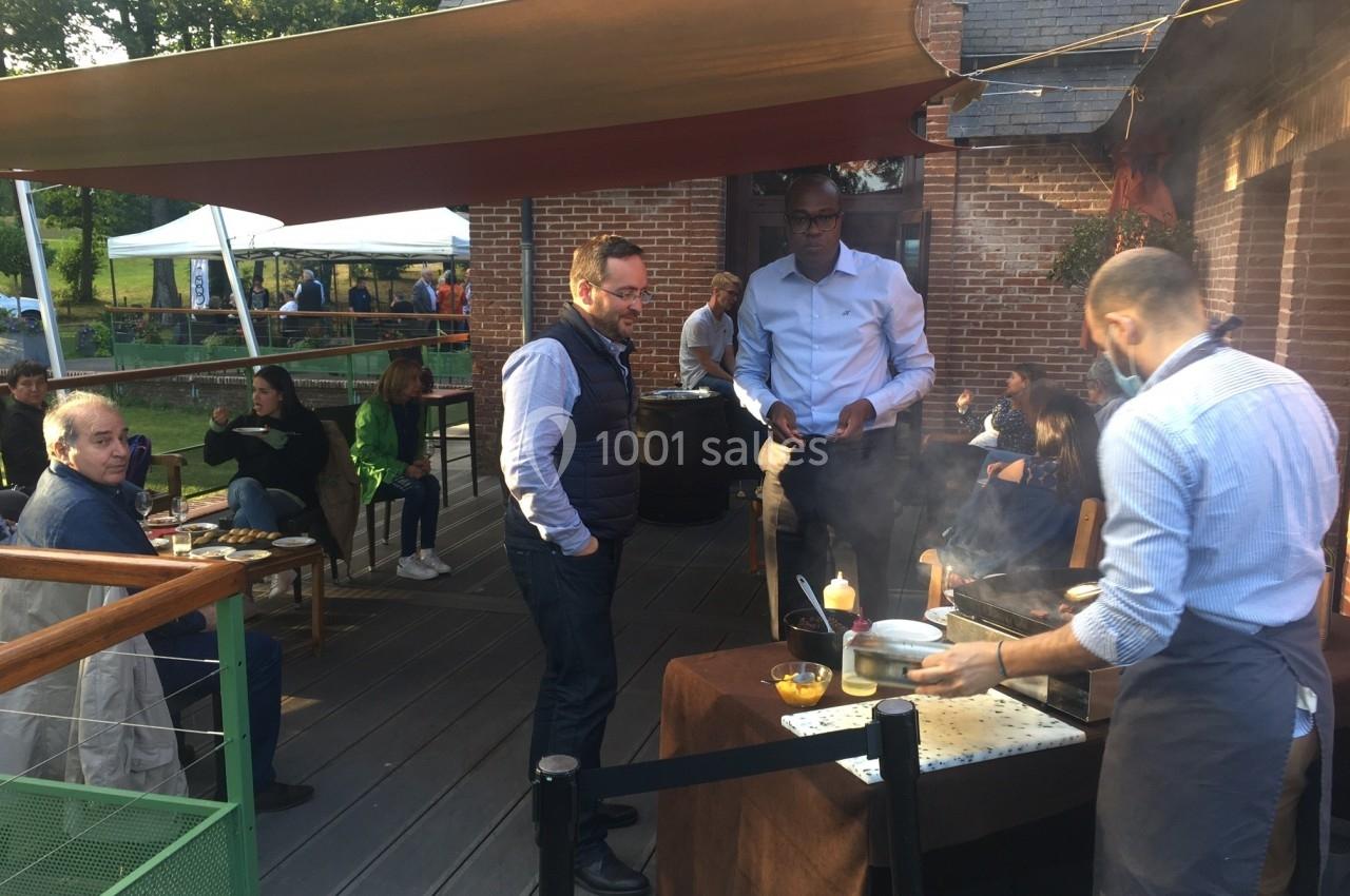 Un cuisinier prépare des plats sur une terrasse en bois, entouré de clients assis et debout dans une ambiance conviviale.