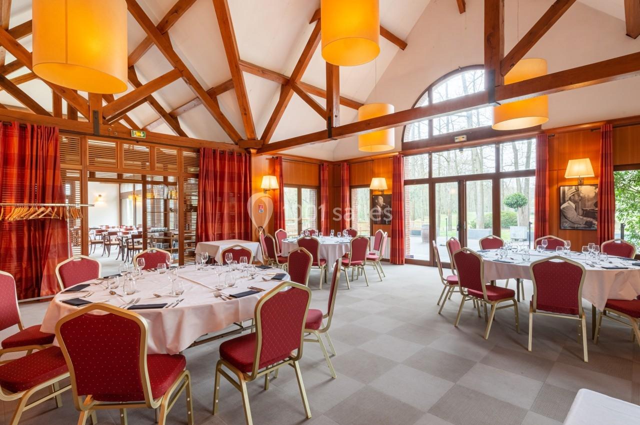 Salle de réception lumineuse avec tables rondes dressées, chaises rouges et grandes baies vitrées donnant sur un jardin.