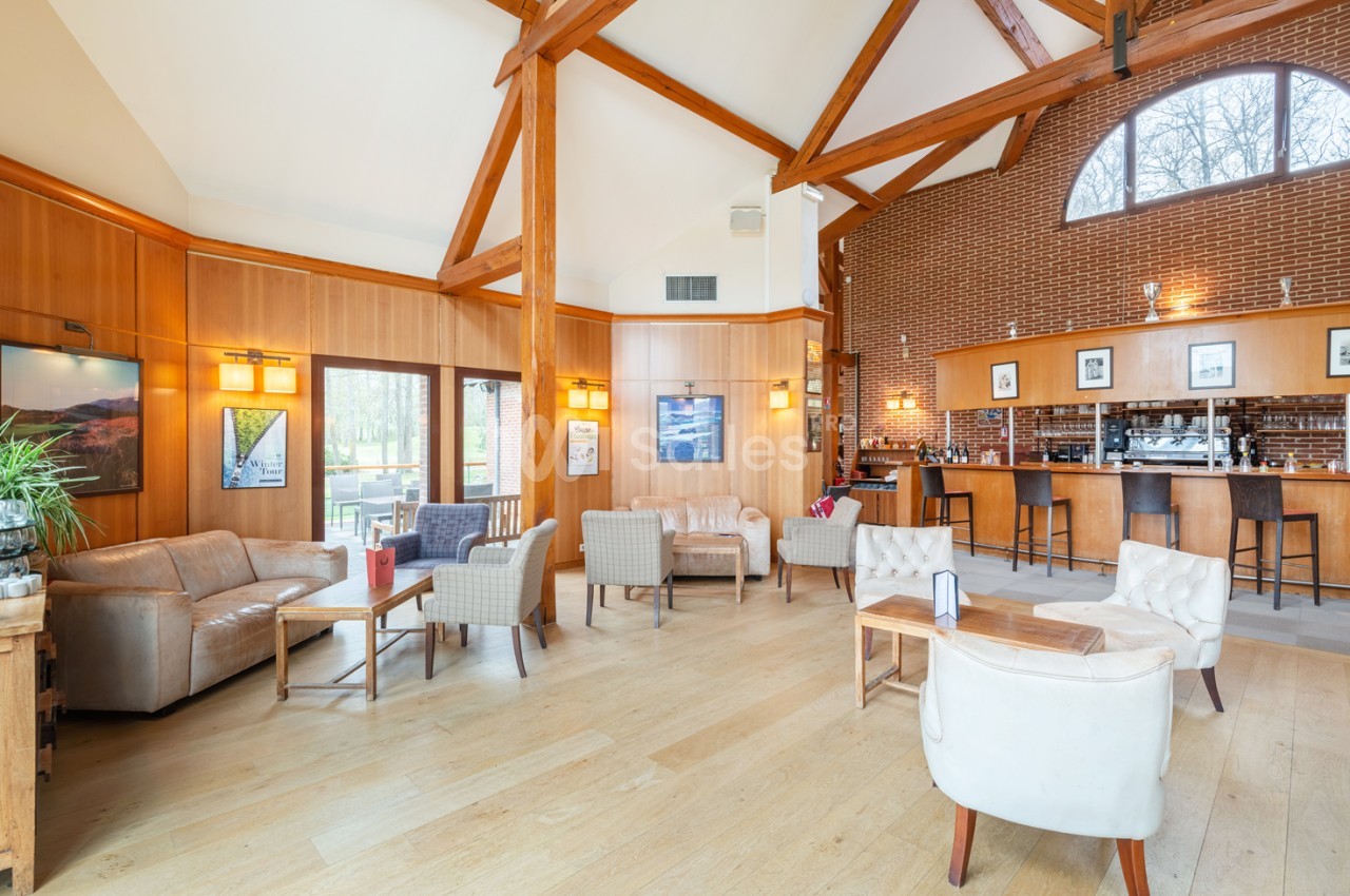 Salon lumineux avec poutres apparentes, mobilier confortable, bar en arrière-plan et grandes fenêtres donnant sur l…