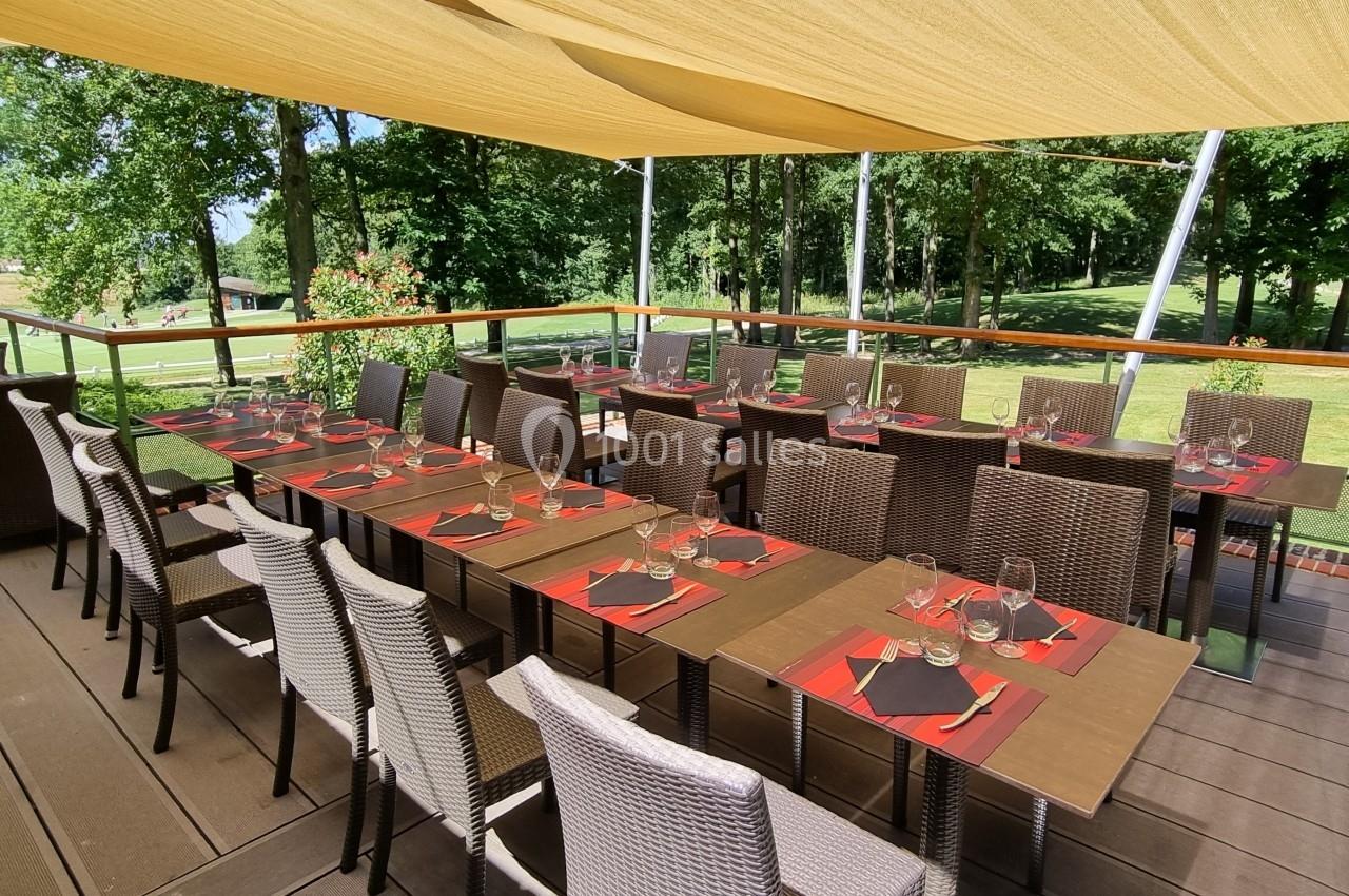 Terrasse ombragée avec tables dressées, chaises en osier et vue sur un espace vert arboré.