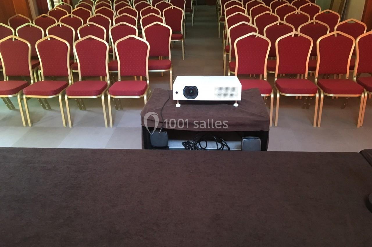 Salle de conférence vide avec des rangées de chaises rouges alignées et un projecteur placé sur une table au premier plan.