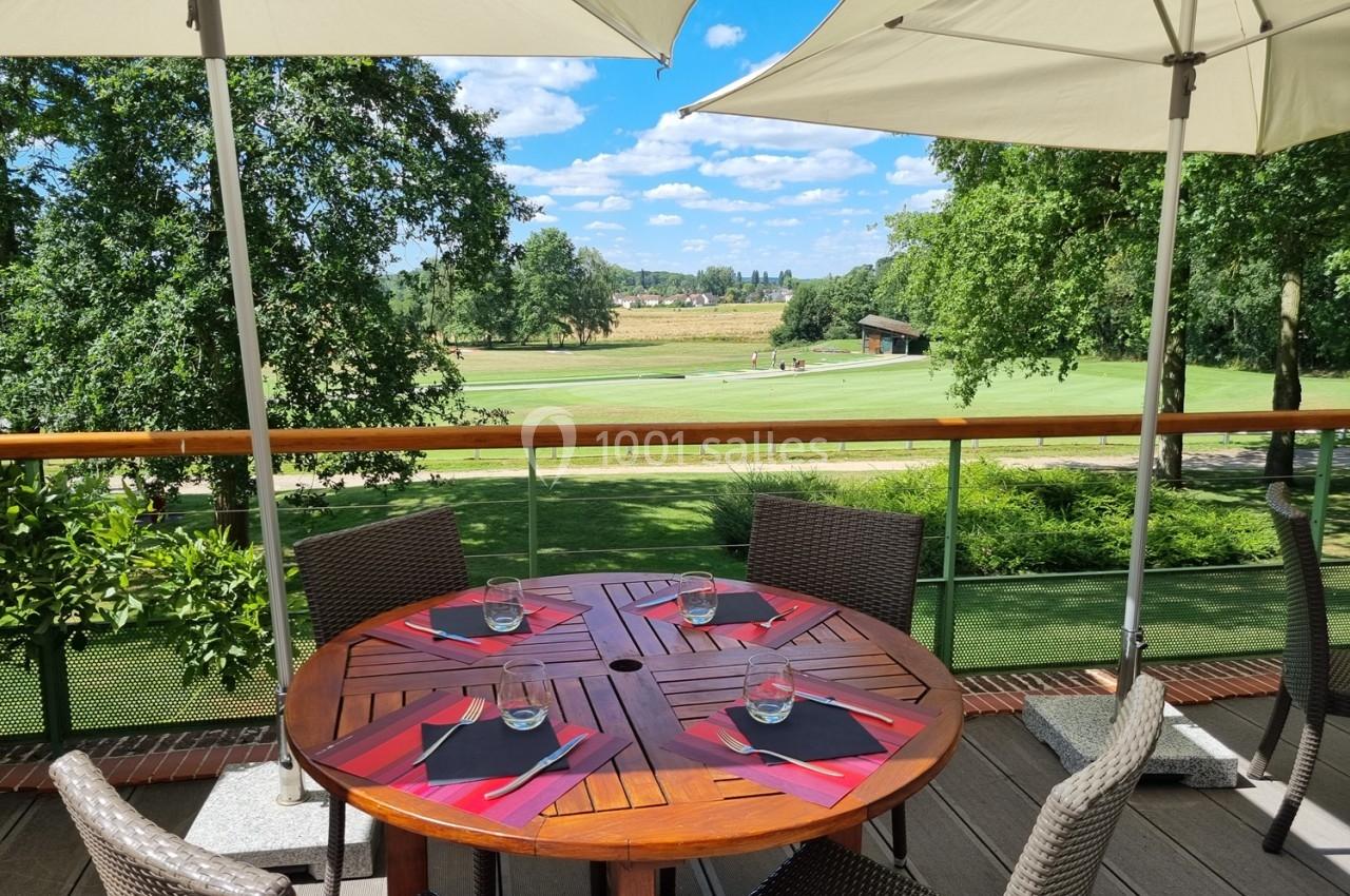 Table en bois dressée avec des sets rouges sur une terrasse ombragée, vue sur un terrain de golf verdoyant.