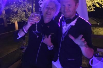 Vue nocturne d'une maison en bois éclairée, entourée d'arbres illuminés, sous un ciel nuageux avec la lune visible.