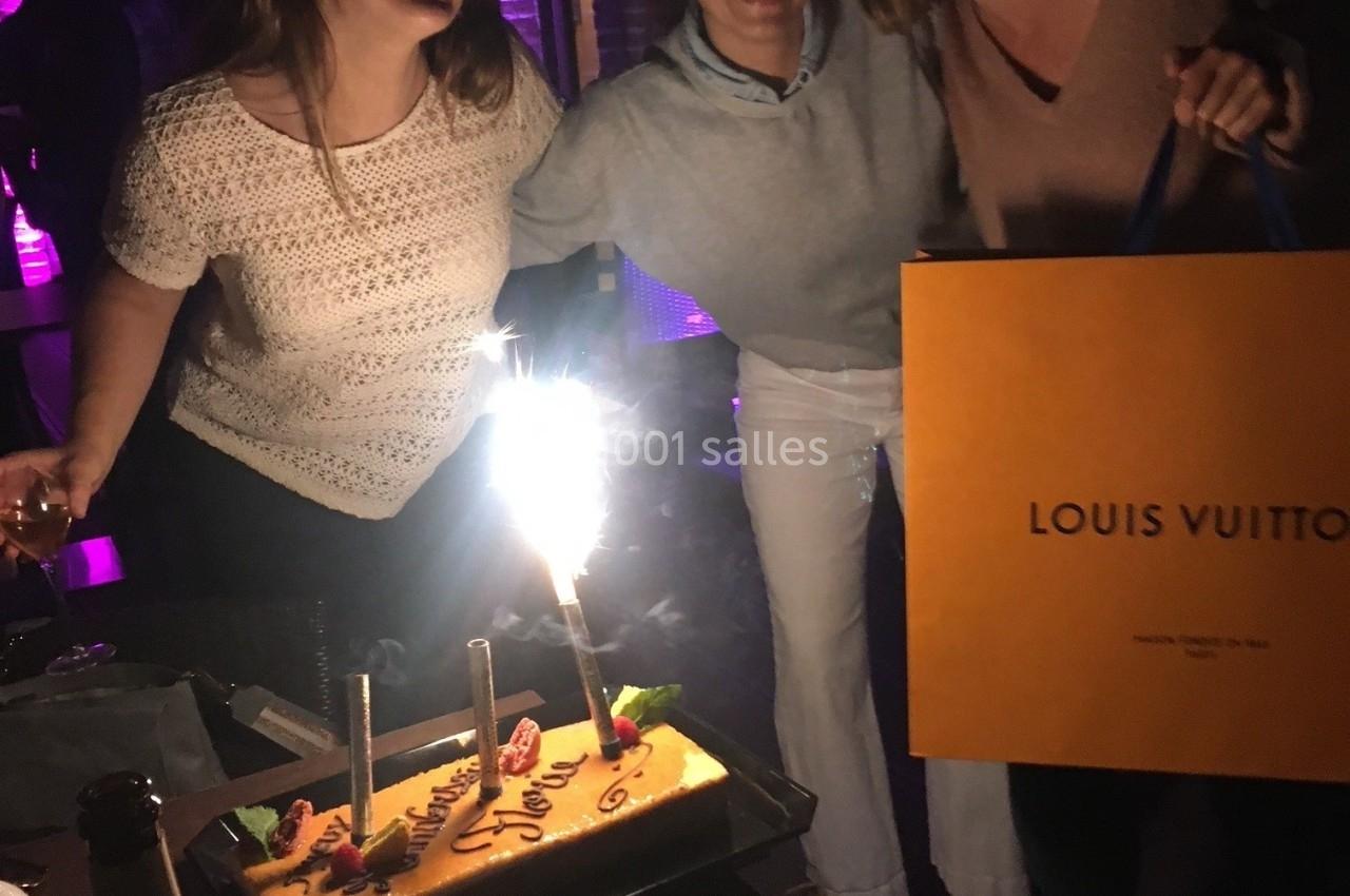 Trois femmes souriantes autour d'un gâteau d'anniversaire éclairé par une bougie étincelante, dans un cadre nocturne.
