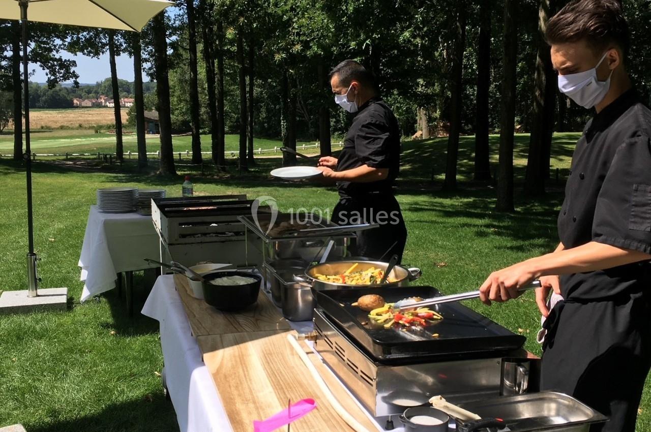 Deux cuisiniers masqués préparent des plats sur des plaques chauffantes lors d'un buffet en plein air, sous des parasols.